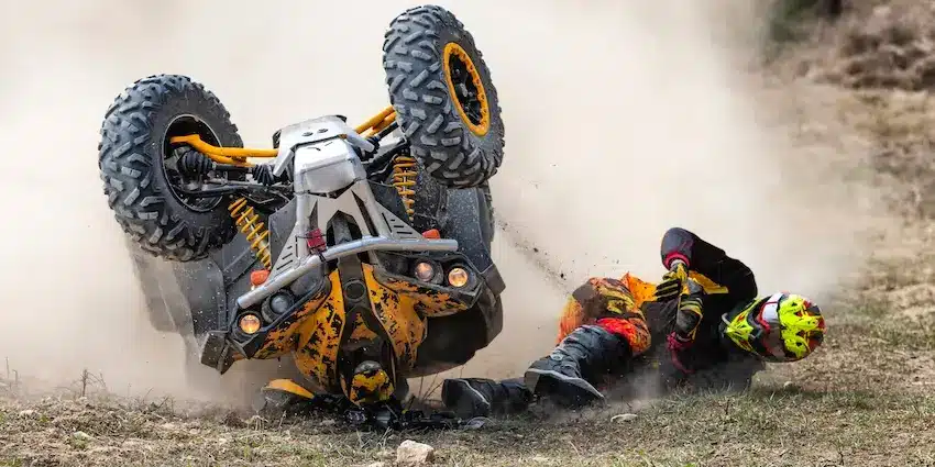 A rider in full protective gear is thrown to the ground as their yellow and gray ATV flips and rolls through a cloud of dust outdoors illustrating the catastrophic injuries that can result when a defective product such as a faulty all-terrain vehicle malfunctions during operation in Georgia.