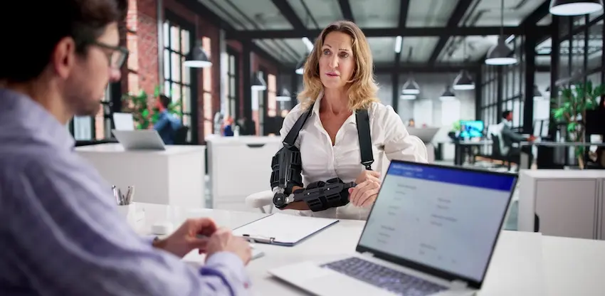 A woman wearing a rigid arm brace leans forward attentively across a desk from a professional with a laptop and clipboard in a modern open office depicting an injured client consulting with an attorney from Hartley Rowe & Fowler P.C. about injuries sustained from a defective product in Georgia.