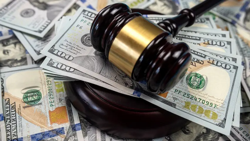 A dark mahogany judge's gavel with a wide shiny gold band rests on its circular sound block which sits directly on top of a large scattered pile of American hundred-dollar bills fanned out across the entire surface of the frame with the green and black printed faces of Benjamin Franklin- serial numbers- and treasury seals clearly visible on multiple overlapping bills beneath the gavel — a powerful visual representation of the monetary compensation awarded to injury victims to cover economic damages- with help from Hartley, Rowe & Fowler P.C.- such as medical bills- lost wages- and property losses in Georgia.
