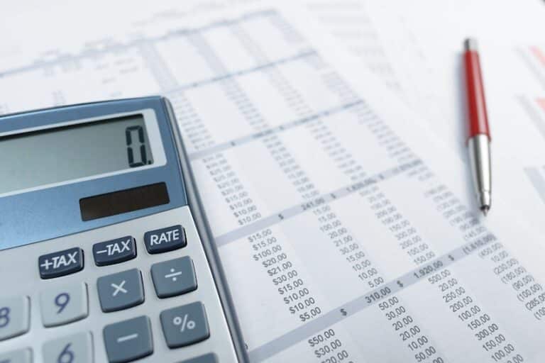 A silver desktop calculator with a digital display reading zero and clearly labeled function keys including plus tax- minus tax- and rate sits on the left side of a printed financial spreadsheet filled with dense columns of dollar amounts- figures- and numerical data with a red and silver ballpoint pen resting diagonally across the upper right portion of the document against a white surface — depicting the detailed financial accounting process used by attorneys from Hartley Rowe & Fowler P.C. to calculate and document economic damages in a personal injury claim.