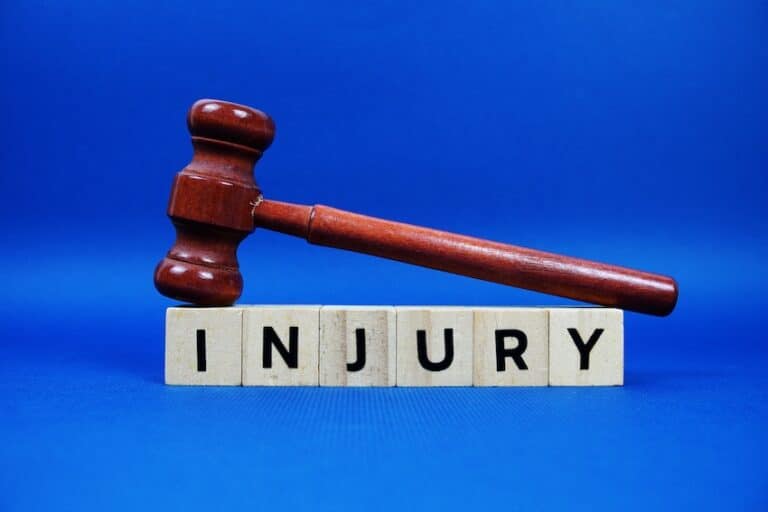 A dark wooden judge's gavel resting diagonally across six wooden letter blocks spelling out the word "INJURY" against a vivid blue background visually representing the legal process of filing injury claims against a negligent party with help from Hartley Rowe & Fowler P.C.