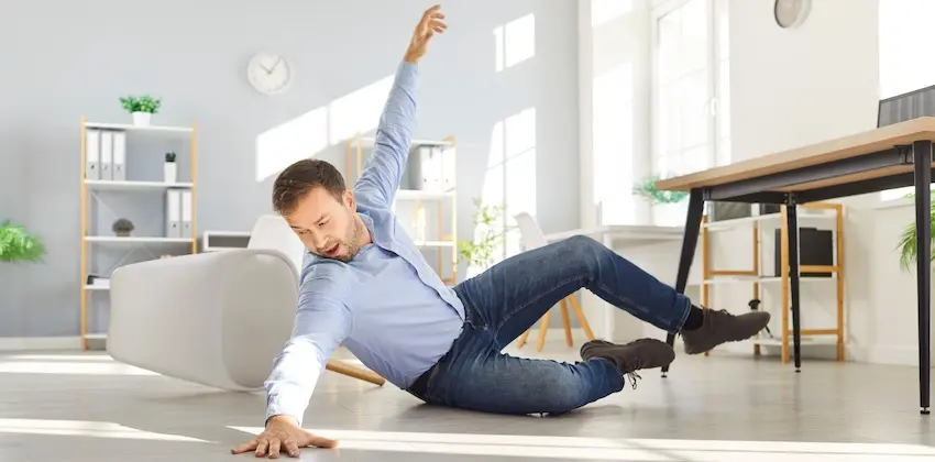 A man with short dark hair and light stubble wearing a light blue button-down shirt and dark jeans has just slipped and fallen on the smooth white floor of a bright modern office space- his right arm raised in the air and left hand braced against the floor as he lands on his side with a pained expression on his face with a gray sofa- open shelving unit with binders and a small potted plant- a round wall clock- and a wooden desk with a monitor visible in the softly lit background — capturing the moment of a workplace slip and fall accident that gives rise to a claim for economic damages including lost income and medical treatment costs in Georgia.