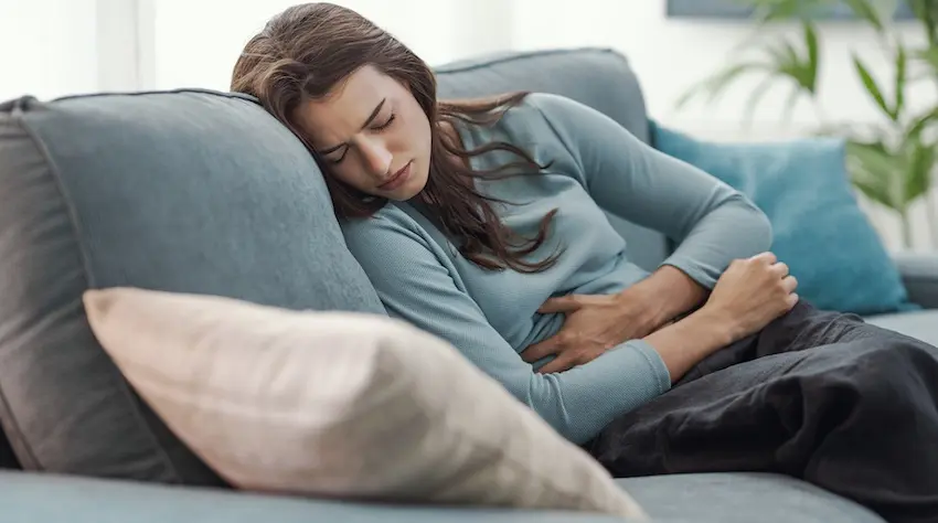 A young woman with long brown hair curled up on a gray couch clutching her abdomen in pain with her eyes closed depicting the kind of ongoing internal pain and physical suffering that drives accident victims to pursue injury claims for compensation in Georgia.