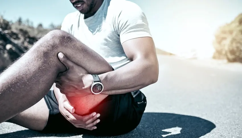A bearded man in a white t-shirt and black shorts sits on a road pavement grimacing and gripping his knee with both hands with a red highlight overlay on the joint indicating the area of acute pain while wearing a sport watch representing the type of documented knee injury that requires thorough medical documentation to establish the full extent of damages in a personal injury claim in Georgia.