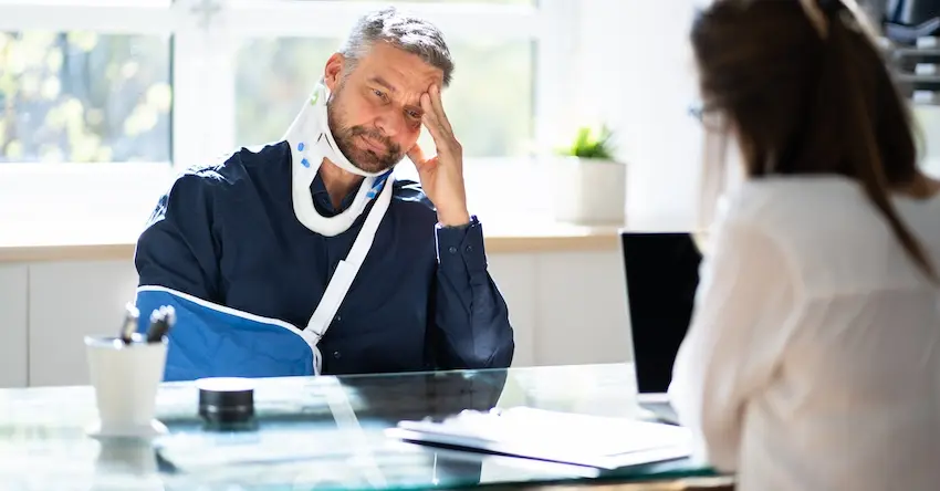 A middle-aged man wearing a cervical neck brace and a blue arm sling sits at a glass desk with his hand pressed to his forehead in visible frustration meeting with a n attorney from Hartley Rowe & Fowler P.C. across the table to discuss the details of his injury claims following a serious accident in Georgia.