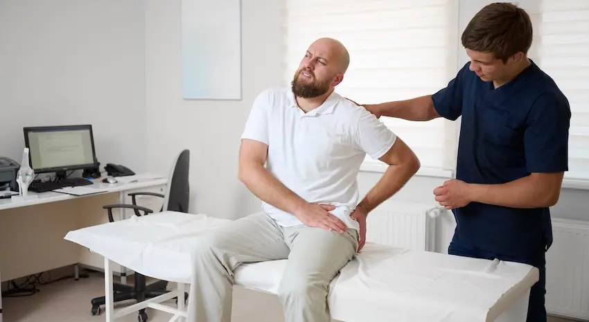 A bald bearded man in a white polo shirt sits on a padded medical exam table grimacing and gripping his lower back while a young male physical therapist in dark navy scrubs stands beside him with a hand on his shoulder illustrating the ongoing chiropractic and rehabilitative care that is central to documenting and supporting injury claims after an accident in Georgia.